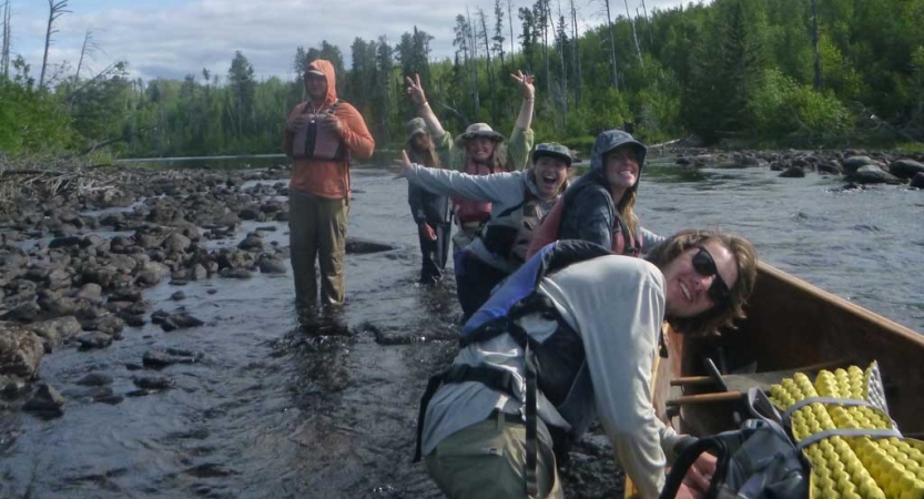 A group of students wearing lifejackets stand in ankle-deep water near a canoe and smile for the photo.